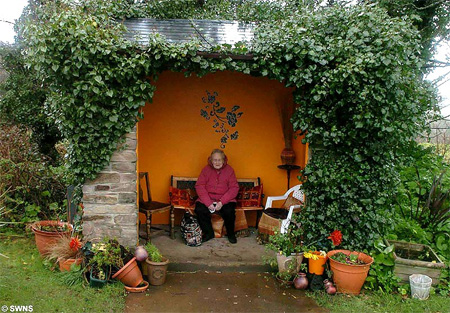 A bus stop adorned with flowers, vines, and potted plants.  A lady sits inside on a cushioned seat.